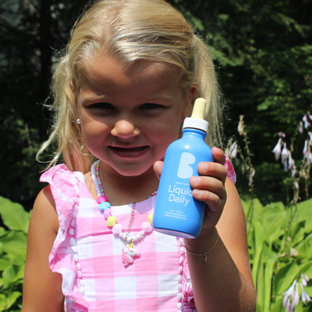 Smiling young girl in a pink dress holding Better Family Liquid Daily vitamin outdoors, showcasing kid-friendly daily nutrition support.