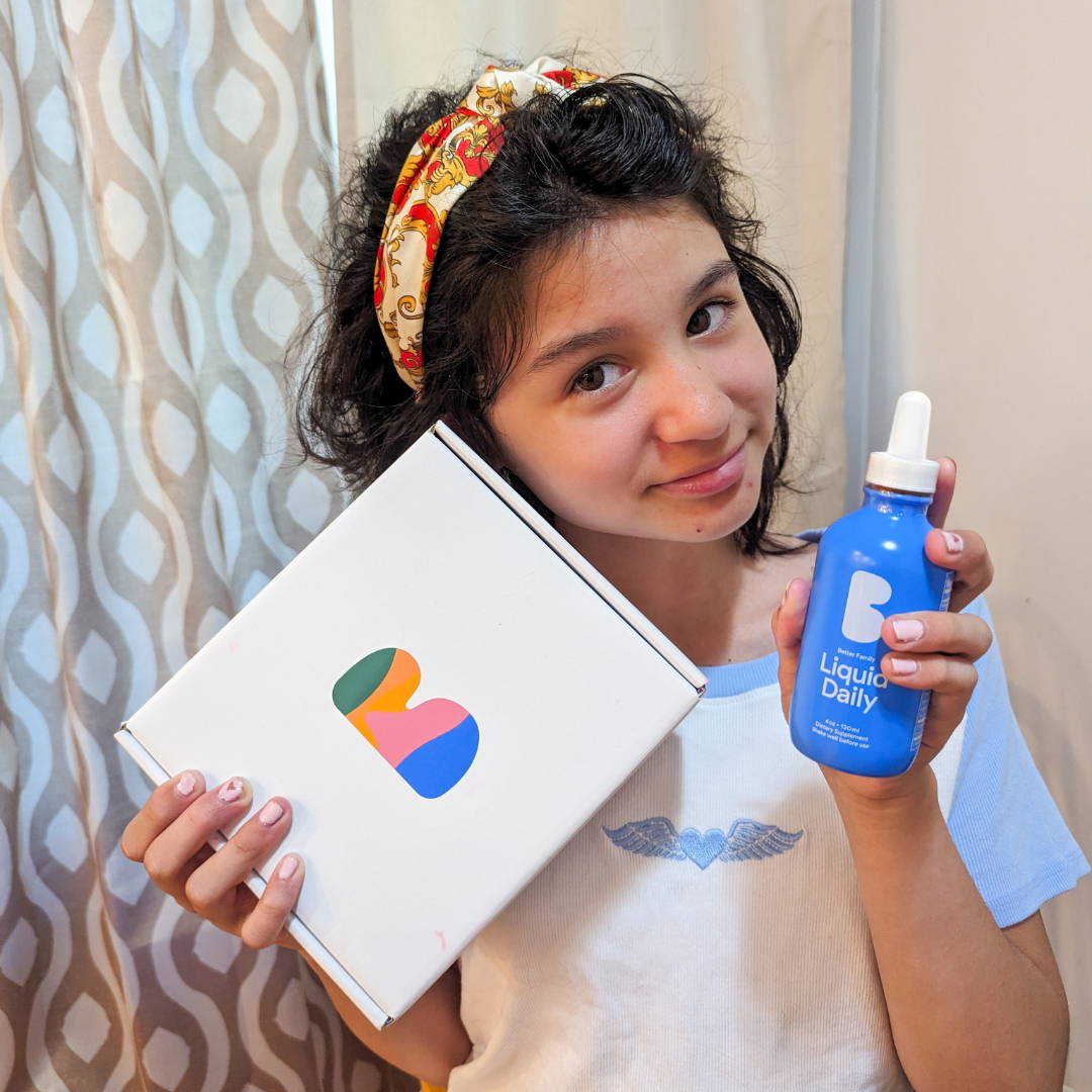 Young girl holding Better Family Liquid Daily supplement and branded box, smiling in a cozy home setting.